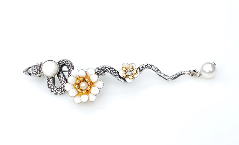 Broche serpent en métal argenté patiné. le long de son corps sont posés un cabochon de nacre blanche, une fleur pompon en émail époxy blanc, une petite fleur en émail blanc et un cristal Swarovski, une perle d'eau douce en pampille au bout de la queue de cet élégant serpent.  Modifier le texte alternatif
