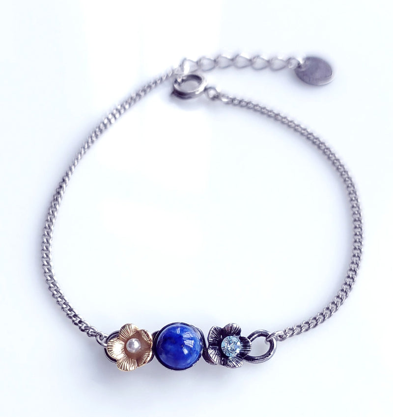délicat bracelet fleuri en métal argenté patiné, réhaussé d'une petite fleur dorée à l'or fin avec une perle blanche en son coeur. Au centre du bracelet un cabochon en pierre fine lapis lazuli et dans la deuxième fleur qui entoure le lapis , un cristal Swarovski. Longueur 16,5 cm environ plus chaine de réglage.