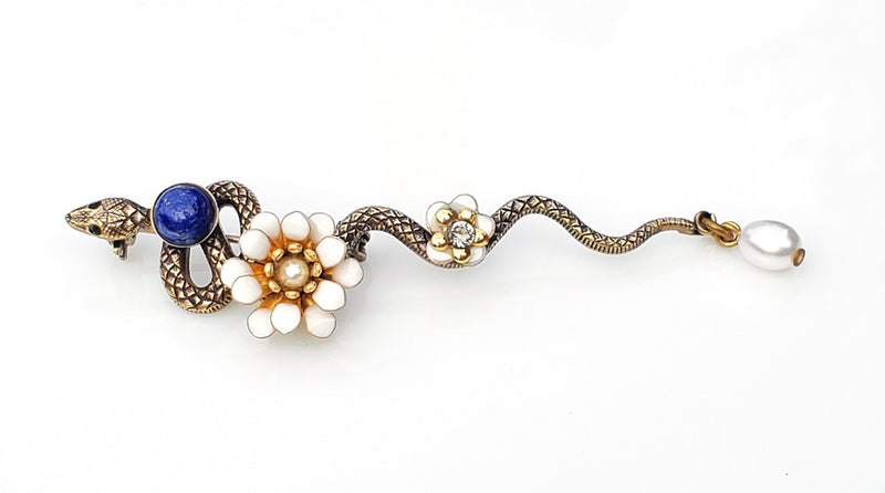 Broche serpent en métal bronze patiné. le long de son corps sont posés un cabochon en lapis lazuli, une fleur pompon en émail époxy blanc, une petite fleur en émail blanc et un cristal Swarovski, une perle d'eau douce blanche en pampille au bout de la queue de cet élégant serpent.  Modifier le texte alternatif