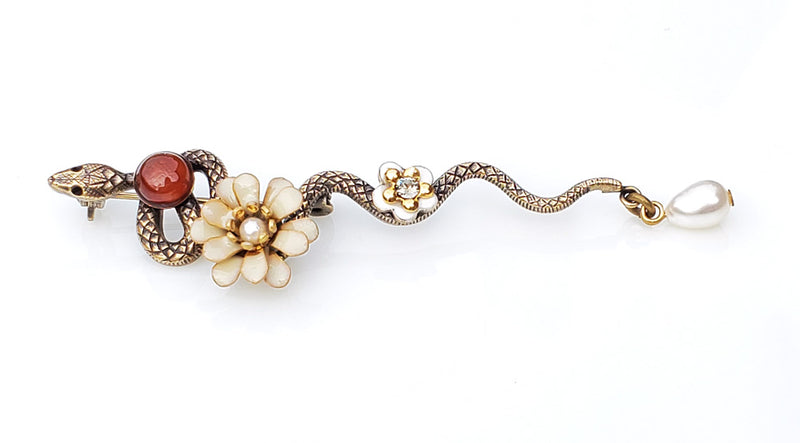 Broche serpent en métal bronze patiné. le long de son corps sont posés un cabochon de pierre cornaline, une fleur pompon en émail époxy crème, une petite fleur en émail blanc et un cristal Swarovski, une perle d'eau douce blanche en pampille au bout de la queue de cet élégant serpent.