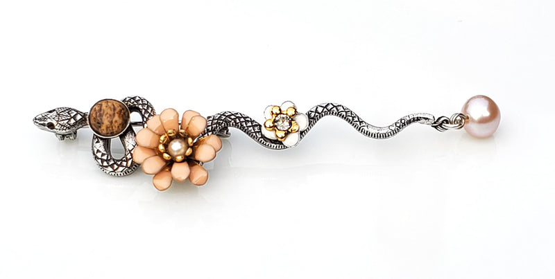Broche serpent en métal argenté patiné. le long de son corps sont posés un cabochon en pierre en jaspe paysage, une fleur pompon en émail époxy rose nude, une petite fleur en émail blanc et un cristal Swarovski, une perle d'eau douce rose en pampille au bout de la queue de cet élégant serpent.