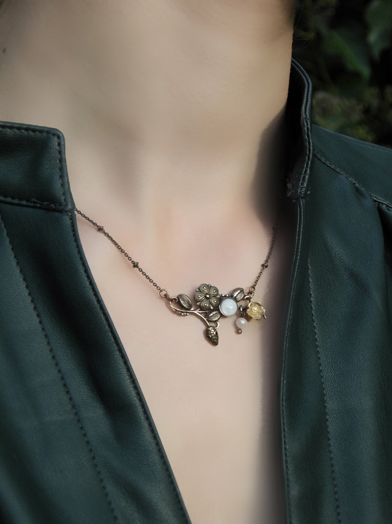 Collier Eden, coloris bronze, un joli tour de cou en métal gravé  d'où émerge une petite tête de serpent parmi  fleurs et feuillage. Pour illuminer la scène, une petite fleur dorée à l'or fin, un cabochon de nacre blanc et une petite perle d'eau douce en pampille.