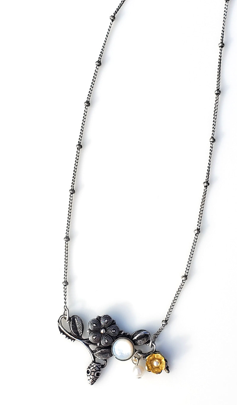 Zoom sur le motif du collier Eden, coloris argent. Une petite tête de serpent émerge parmi les fleurs et les feuilles. la pureté du métal est soulignée par la blancheur de la nacre, perle et cabochon.