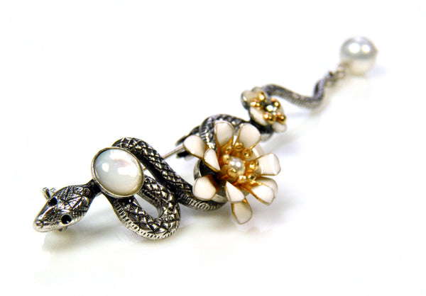 Broche serpent en métal argenté patiné. le long de son corps sont posés un cabochon de nacre blanche, une fleur pompon en émail époxy blanc, une petite fleur en émail blanc et un cristal Swarovski, une perle d'eau douce en pampille au bout de la queue de cet élégant serpent.
